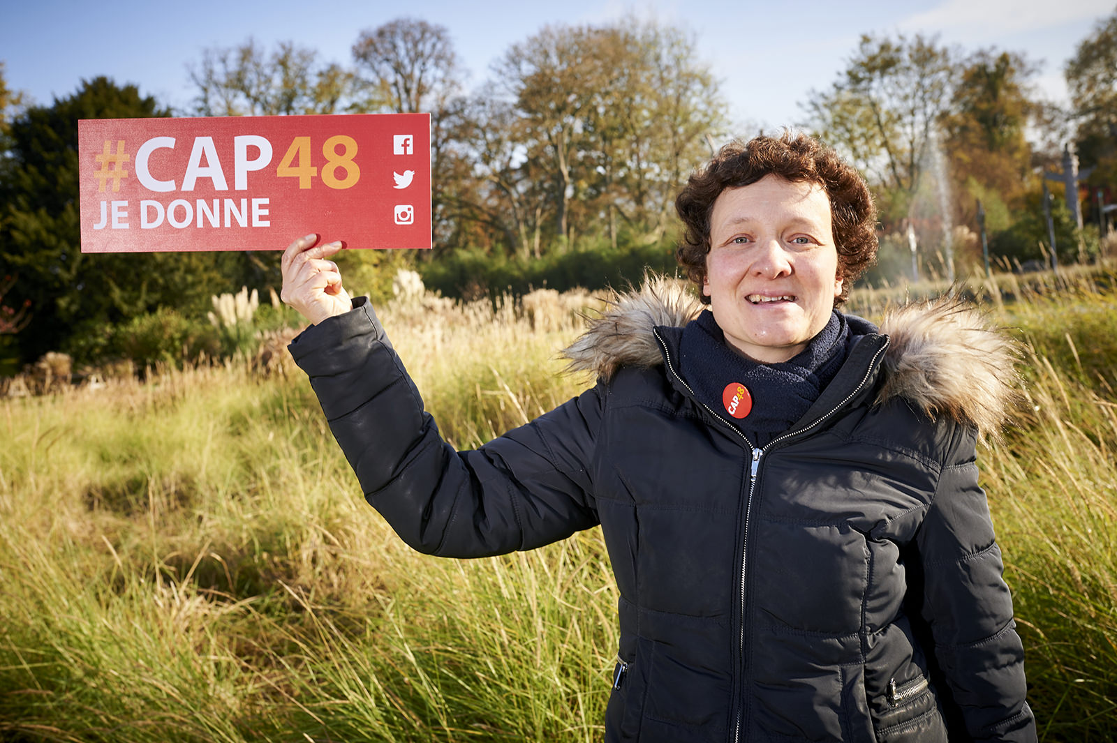 Une femme, bénévole CAP48, souriant et tenant en main une pancarte avec le slogan : "CAP48 je donne" lors d'une campagne d'appel aux dons