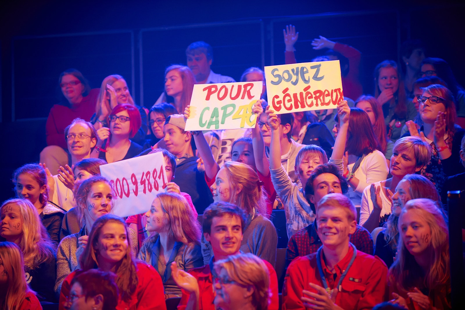 Public de spectateurs lors de la Grande Soirée CAP48 brandissant des pancartes d'appels à faire un don